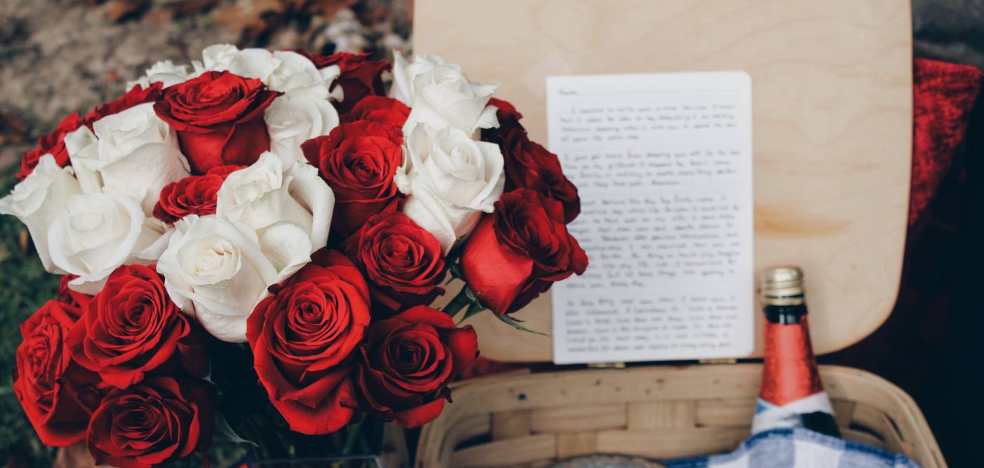 A bouquet of red and right roses next to a basket with a handwritten note and a bottle of sparkling wine inside